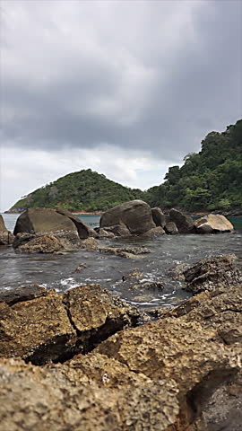 Natural landscape with rocks, water on a calm beach, and lush hills in the background.