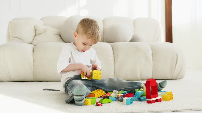 Young child focuses on assembling colorful building blocks while seated on a soft carpet in a well-lit living room with a modern sofa