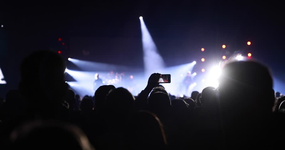 Close-up of a person holding a mobile phone to record a band on stage with intense white and red flashing spotlights and strobe effects.