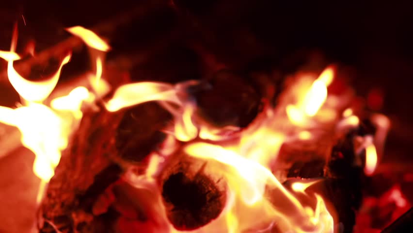 A close view of a roaring fire with bright orange and yellow flames. The wood logs are glowing and turning into charcoal under intense heat.
