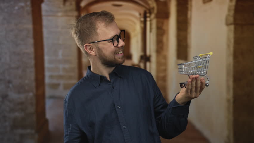 Young caucasian man smiling while holding a miniature shopping cart on his open palm in a building corridor; consumer optimism.