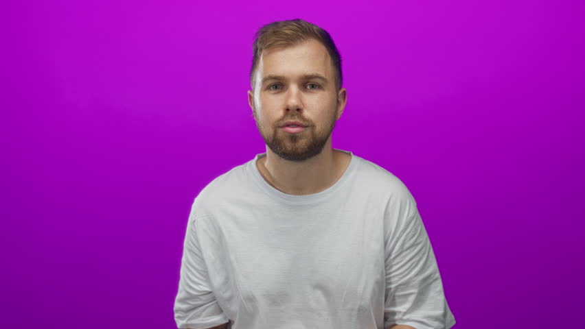Young caucasian man smiling with hands giving thumbs up and clenched fist gesture in purple studio wearing white t shirt; excitement celebration energy.