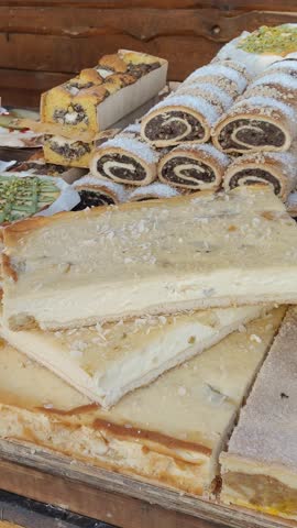 Assorted homemade cakes and pastries on wooden market stall, creamy layered cheesecake slices and poppy seed rolls with powdered sugar.