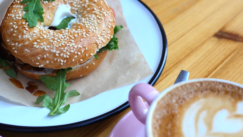 Close up of a sweet breakfast or brunch on a wooden table, featuring a pink cup of cappuccino with latte art, a box of colorful donuts, a bagel sandwich, and sunglasses in a cozy cafe