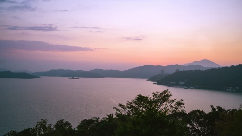 A serene sunset view over the tranquil waters of Sun Moon Lake in Nantou, Taiwan, featuring silhouettes of mountains and lush trees.
