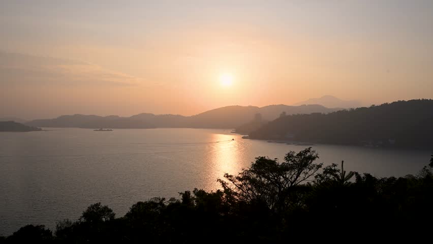 A scenic view of the sun setting over Sun Moon Lake in Nantou, Taiwan, with golden light reflecting on the water and silhouettes of mountains.