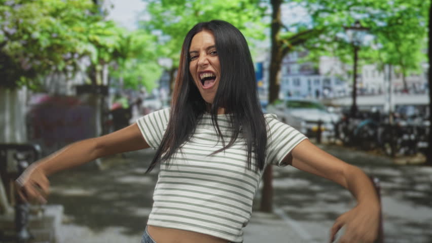 Woman flexing biceps with fists clenched, striped cropped top showing midriff on a leafy urban street with parked bikes; confidence empowerment.