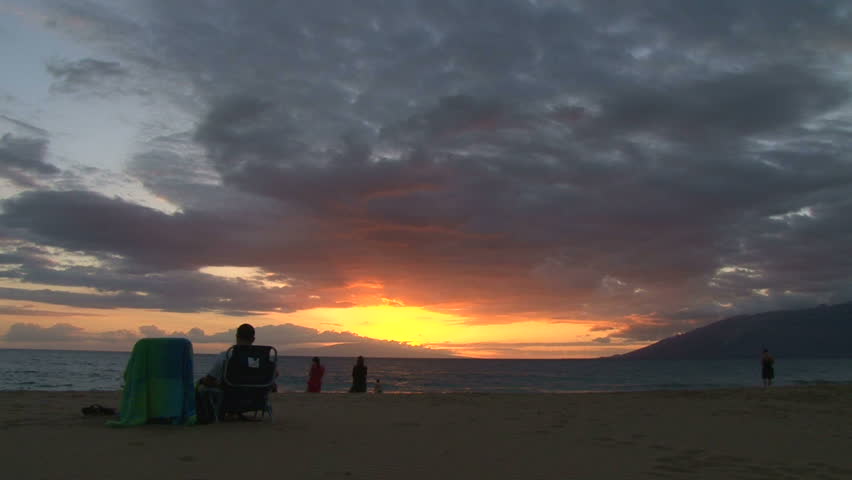 Silhouetted woman and man watch sun go down in Hawaii on sandy beach, real time.