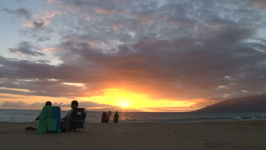 Silhouetted people watching sun go down in Hawaii on sandy beach, time lapse.