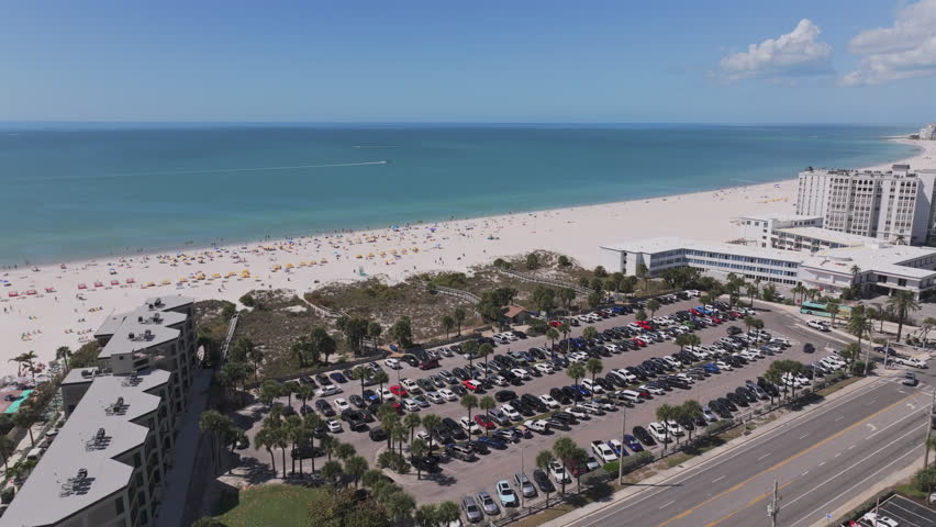 Aerial view of Saint Pete Beach public parking lot packed with cars, beach access boardwalk, beachgoers with umbrellas, and stunning turquoise Gulf waters in Florida