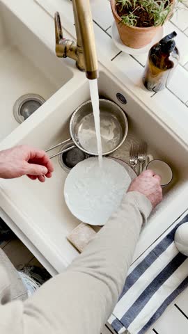 Man washes dishes in a kitchen sink with a sponge and liquid soap under running water. Contemporary domestic scene and modern masculinity. Concept of sharing household chores. Day light, close-up