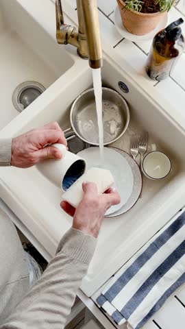 Man washes dishes in a kitchen sink with a sponge and liquid soap under running water. Contemporary domestic scene and modern masculinity. Concept of sharing household chores. Day light, close-up