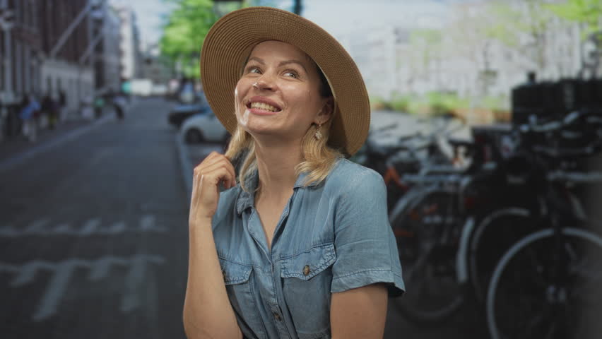 Blonde woman wearing straw hat and denim shirt smiles and touches hair on sunlit cobblestone street; carefree happiness.