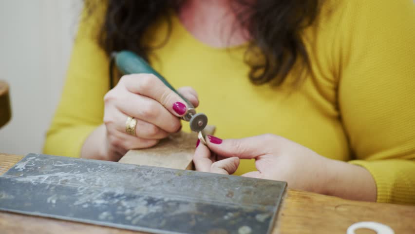 Close up of a female jewelry maker's hands using a small rotary tool to polish and sand a silver metal piece, working at a traditional wooden workbench in her artisan studio