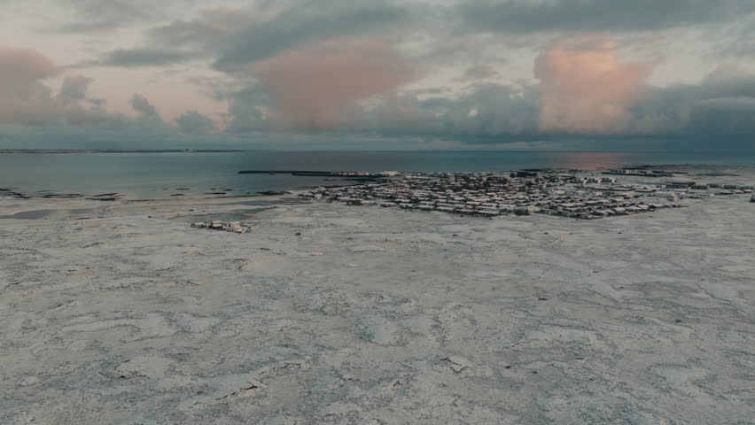 Drone aerial view of a small coastal town in Iceland covered in snow during winter. Remote Nordic settlement surrounded by frozen terrain, ocean and dramatic sky at sunset.