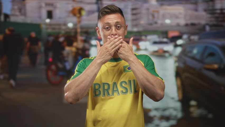 Man in brasil jersey covers mouth with hands on a busy street at night with cars and crowd nearby; surprise support celebration.