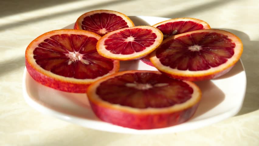 Close-up of fresh blood orange slices arranged on a white ceramic plate. The scene is illuminated by bright, natural sunlight, creating deep shadows and highlighting the rich ruby-red texture of the citrus fruit 4k 
