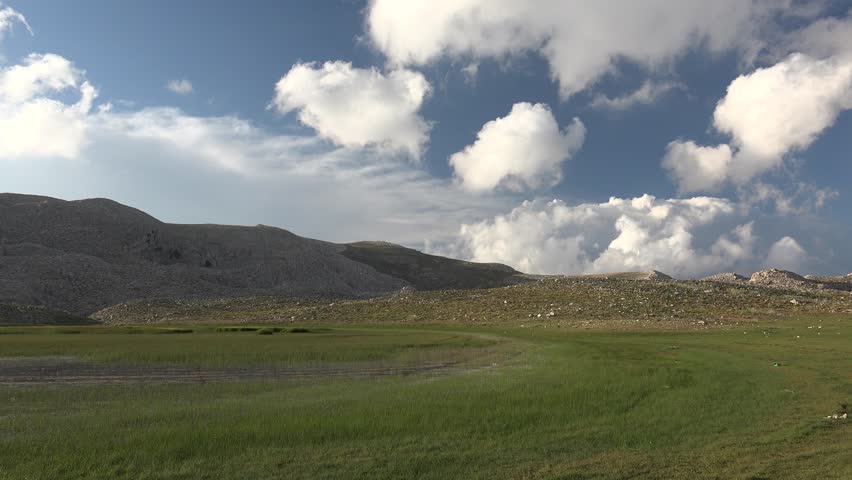 Sunny partly cloudy sky above green plateau pasture and treeless stony hills in open highlands. Timelapse panorama shows summer grass fields, broad tableland, bright clouds, quiet upland scenery.