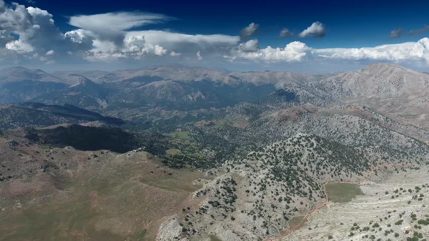 Panoramic aerial view of dirt mountain road crossing green plateau between rugged high peaks. Wide highland landscape reveals winding trail through grassy upland amid remote summits and open terrain.