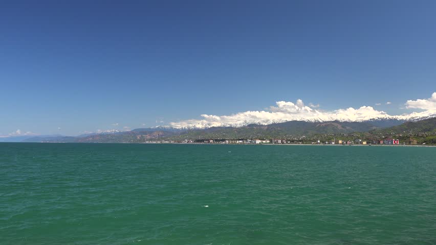 Panoramic coastal settlement beside green forests and snowy mountains under a clear summer sky. Seaside village meets alpine peaks, bright shoreline scenery, sunlit water, woodland, calm bay.