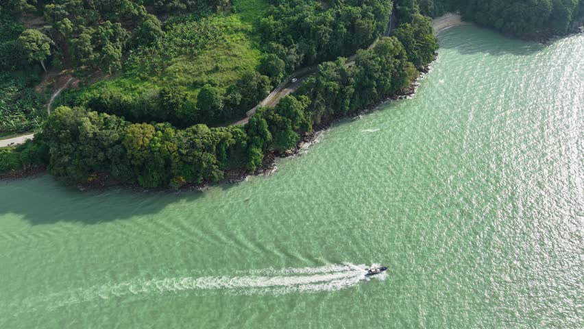View from sky drone view of Balik Pulau coastline, forested promontory, winding road and motorboat leaving wake.