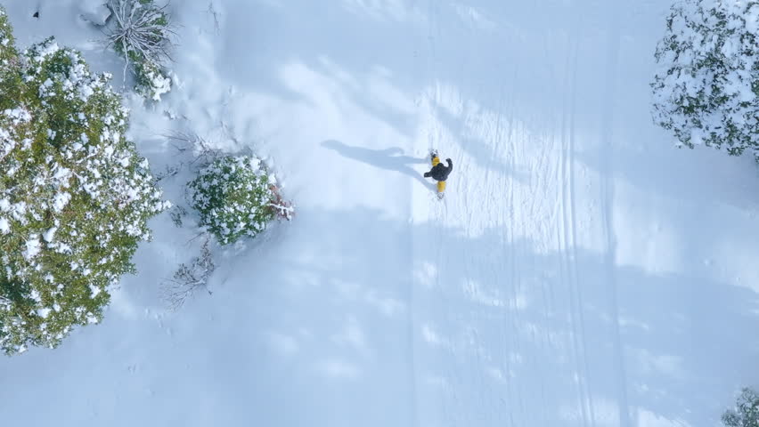 Drone top view capturing a single person walking through deep fresh snow surrounded by winter landscape. Minimal white scenery with peaceful cold atmosphere and clear footprints.