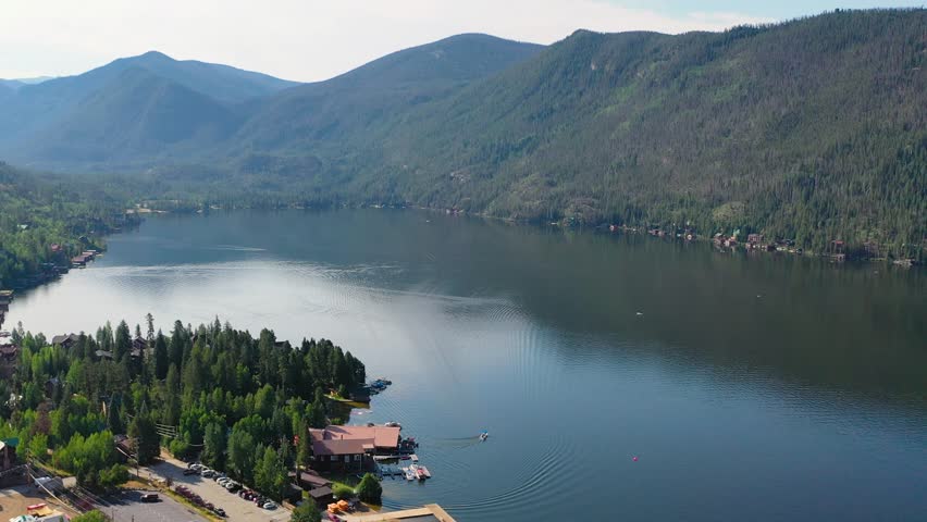 Aerial panorama of Grand Lake Colorado featuring the waterfront downtown, mountain valley, lakeshore homes, boats, and evergreen covered hills under soft summer sunlight