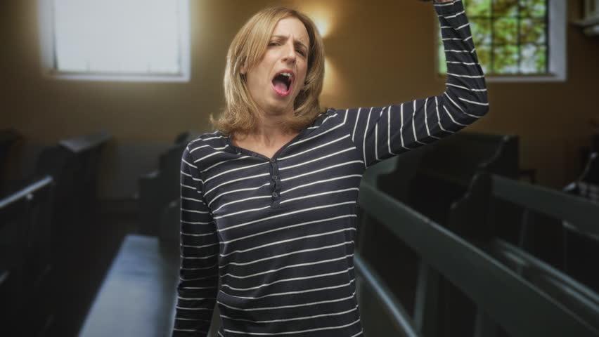 Woman flexes arm and kisses bicep inside building with pews and stained window, puckering lips toward muscle; confidence empowerment strength.