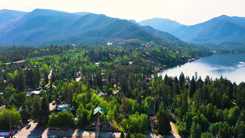 Aerial view of a forested mountain lake community with cabins, shoreline homes, calm water, and layered alpine peaks rising beyond dense evergreen trees in bright summer light