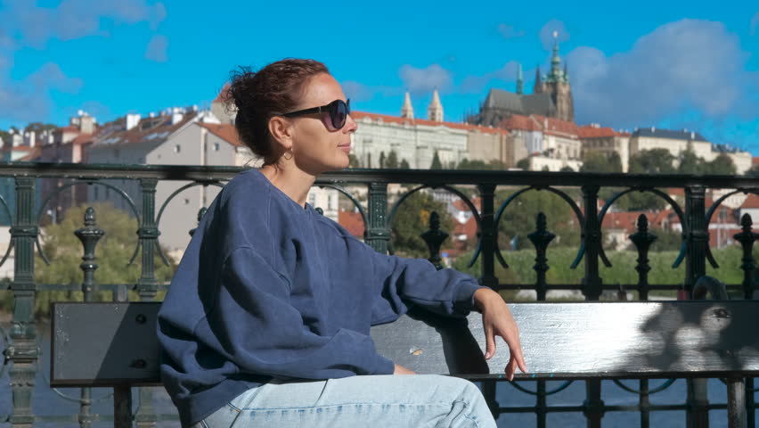 Tourist enjoying prague castle view. Female traveler relaxing on bench, wearing sunglasses and casual attire, overlooking panoramic view of prague castle and vltava river under bright sunny sky