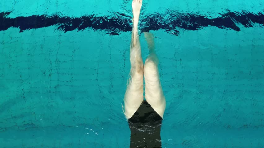Female athlete in a sleek black swimsuit is seen from above as she propels herself off the pool wall to start her backstroke in a crystal clear blue pool