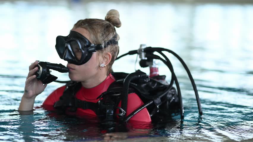 Female diver with scuba gear and mask putting regulator in her mouth and sinking underwater during a certification course in a pool