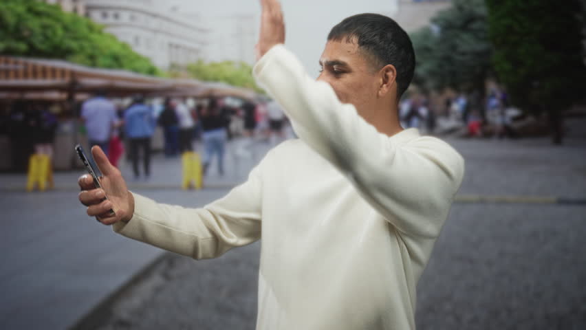 Man holding smartphone, smiling and gesturing with hand on a busy street market plaza crowded with stalls and pedestrians; connection joy.