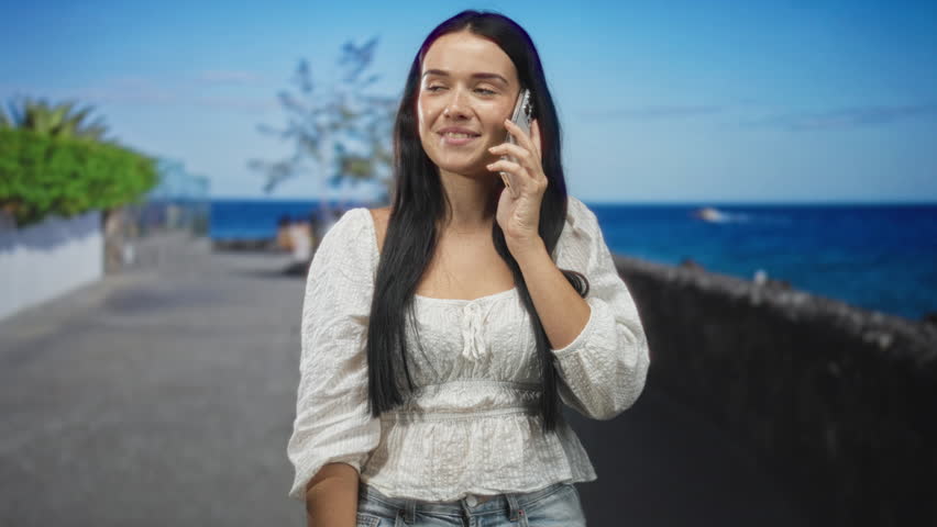 Woman holding smartphone to ear and smiling on a seaside street promenade by the ocean with glass railing and plants visible; serenity.
