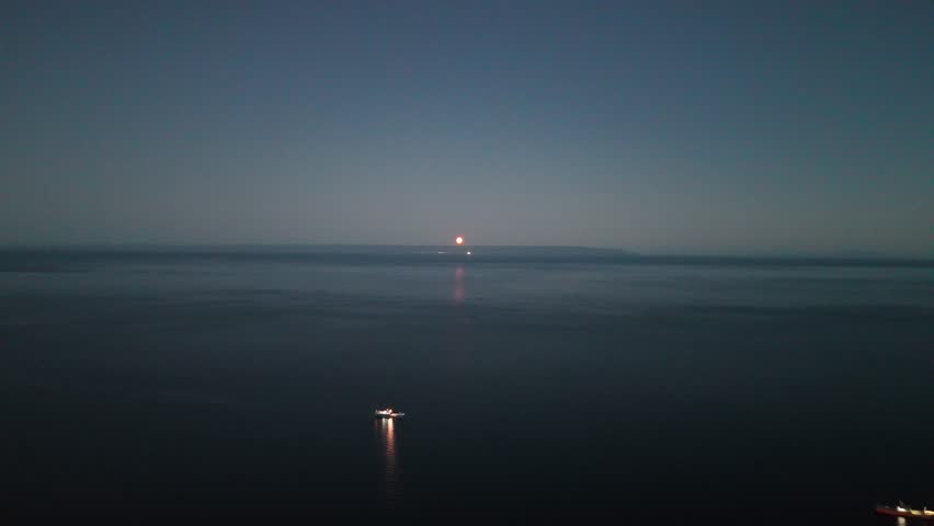 Aerial pulls back from a close-up of the red-hued "Blood Moon" reflecting on the calm waters of the Strait of Magellan, revealing the dark coastline and ships anchored near Punta Arenas, Chile