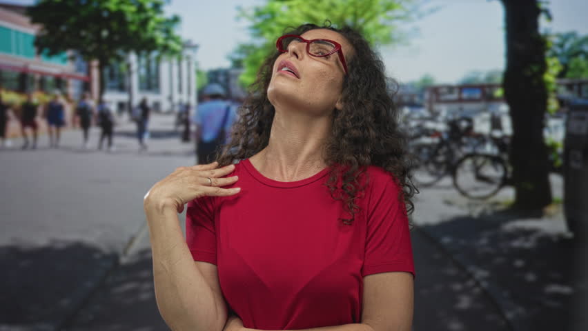 Woman with red glasses and curly hair wearing red shirt raises hand in city street while frowning; annoyance.