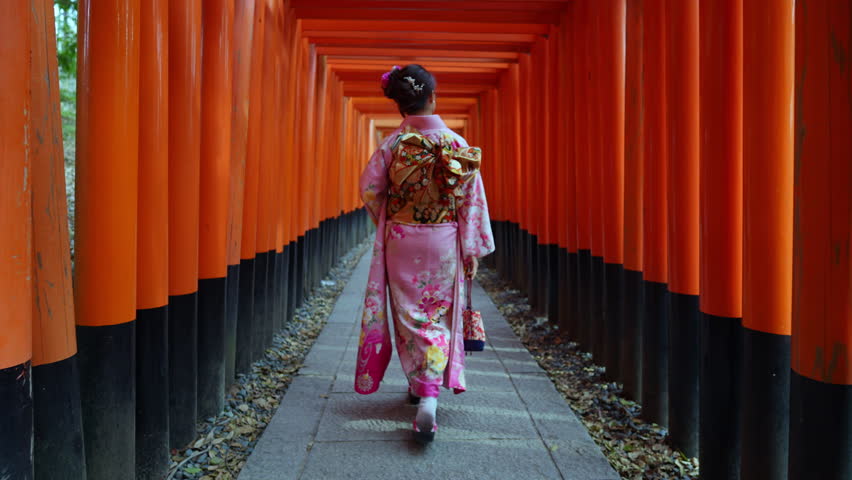 Slow motion video,Asian woman wearing traditional kimono walking through the famous red torii gates at Fushimi Inari Shrine in Kyoto, Japan. Cultural travel destination showcasing historic Japanese heritage and Shinto architecture.