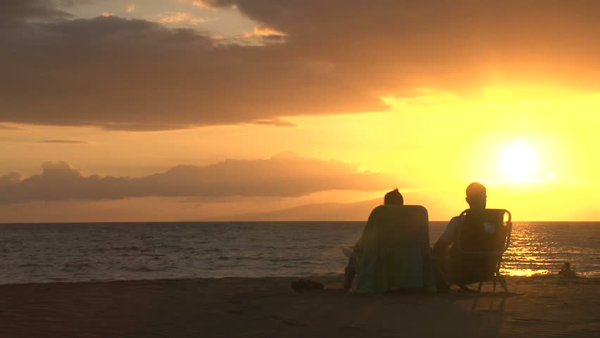 Silhouetted woman and man watch sun go down in Hawaii, sitting on beach chairs