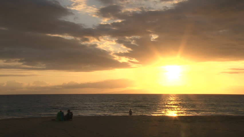 Silhouetted people watch sun go down in Hawaii on sandy beach.