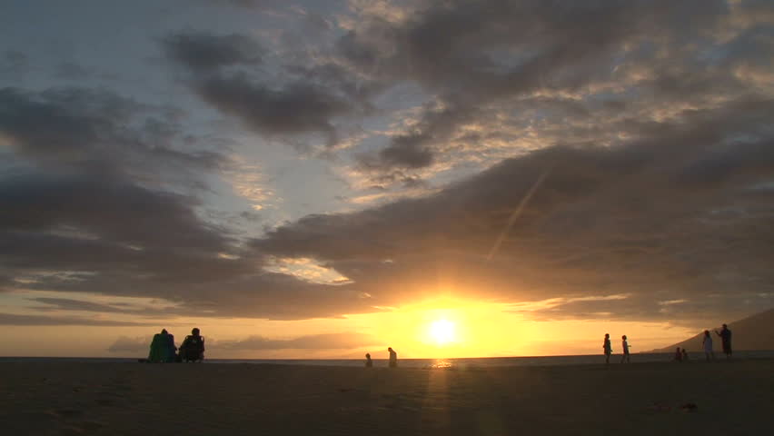 Silhouetted people enjoying sunset in Hawaii on sandy beach.
