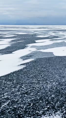 Moving across a frozen sea toward distant ice fishing holes, with dark granular ice channels between white snow sheets under a blue-grey sky, minimalist winter travel shot, vertical.