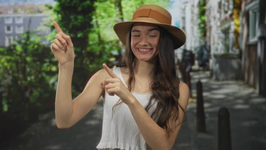 Young hispanic woman pointing finger upwards at something on a sunlit city street while smiling softly; happiness.