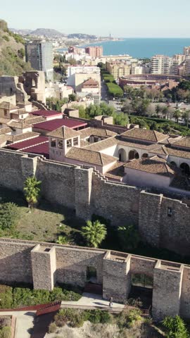 Vertical aerial orbit capturing the historic Gibralfaro Castle walls along the Mediterranean coastline in Málaga, Spain.