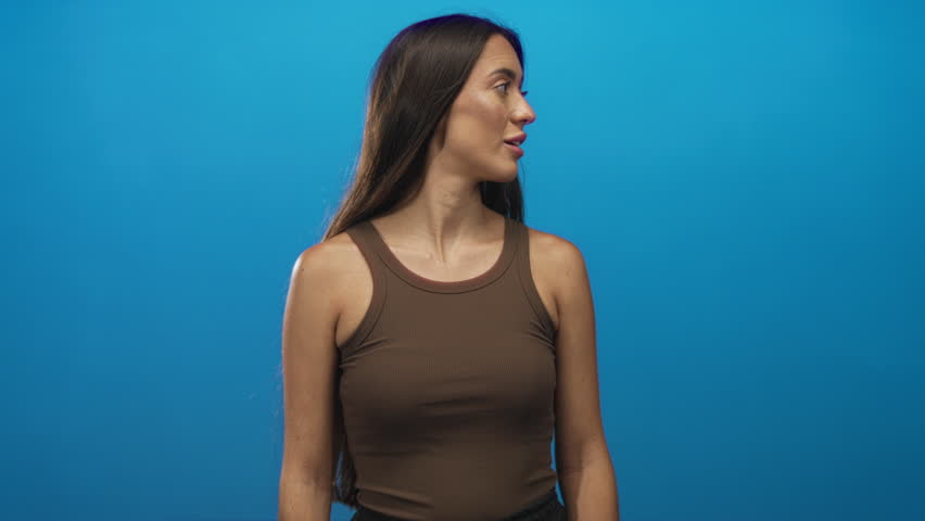 Woman turns head to right in blue studio wearing brown tank top and visible shoulders; contemplation.