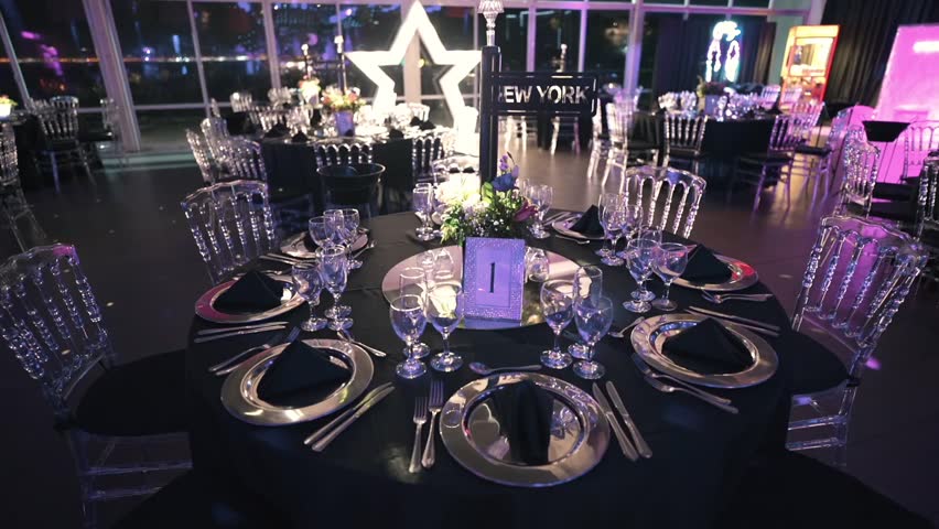 Luxury wedding or gala reception table arrangement with crystal chairs, black tablecloth, silver plates and formal dinner place settings inside illuminated event venue.