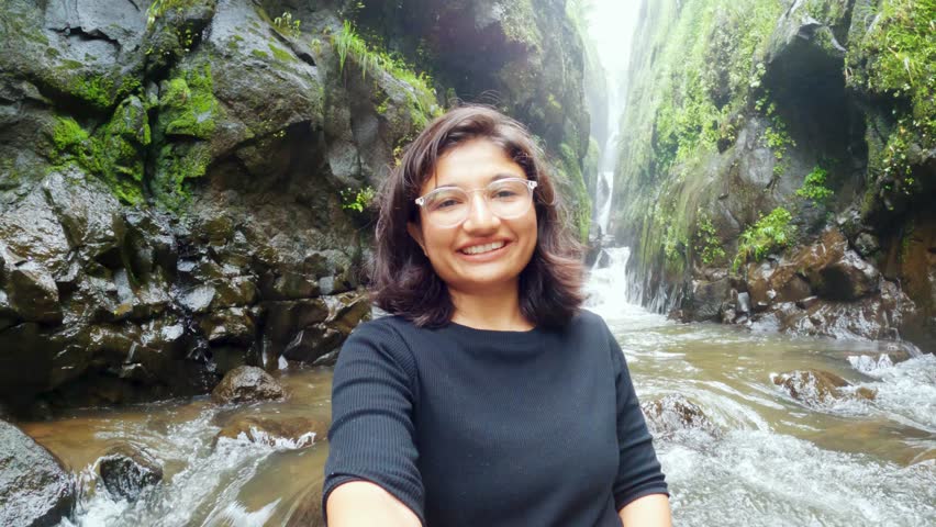 4K Selfie video of Indian teenager girl sitting in front of Satsada waterfall in narrow valley, river stream flowing besides girl. Waterfall background, monsoon landscape. Female tourist recording vlog in mobile phone. Travel, holidays and adventure concept.