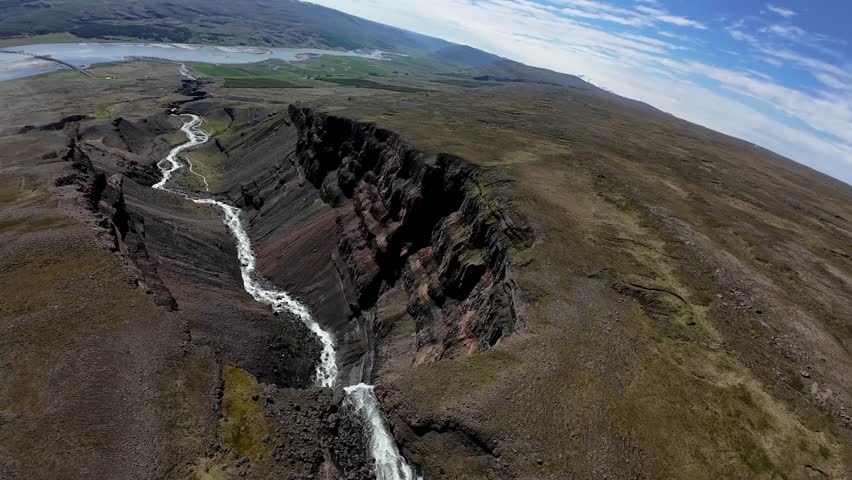 FPV drone glides down a rugged canyon above Hengifoss waterfall near Egilsstaðir, Iceland, passing layered basalt cliffs and misty drops in a fast cinematic summer aerial.