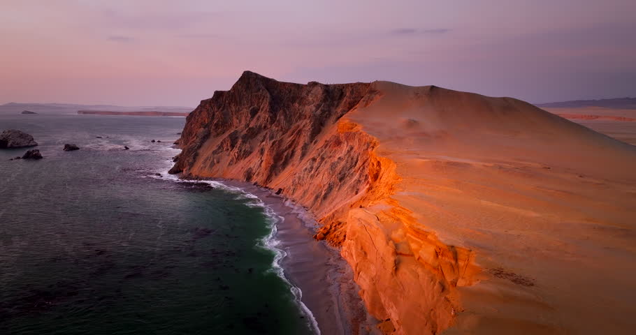 Drone tilts downward over glowing coastal cliffs and beach in Paracas National Reserve, Peru, revealing dark Pacific water and warm desert tones at dusk on South America’s coast