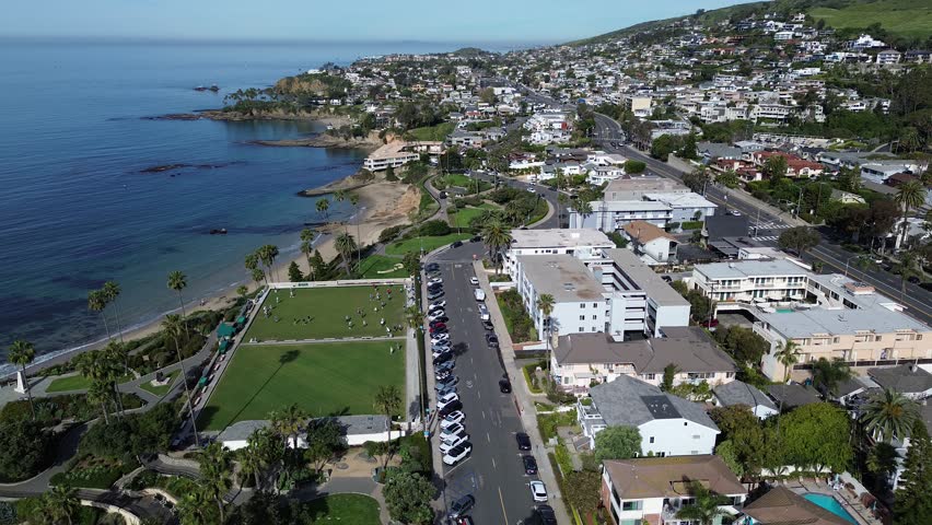 Coastal lawn gathering at Heisler Park near Cliff Drive, Rockpile Beach with visitors spread across the bluff. Manicured trimmed grass, palm shadows, parked cars, mixed roofing, Laguna Beach, CA. USA