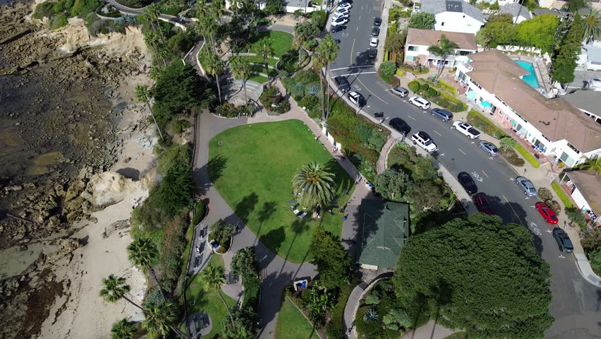 Bluffside park lawn beside Heisler Park and Cliff Drive overlooking Rockpile Beach with visitors near central palm. Bright grass, coastal paths, tree canopy, mixed roofing in Laguna Beach, CA. USA